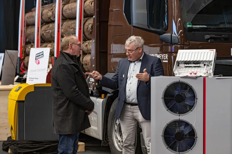 Zwei M&auml;nner im Gespr&auml;ch, im Vordergrund steht eine W&auml;rmepumpe im Hintergrund ein LKW mit Baumst&auml;mmen